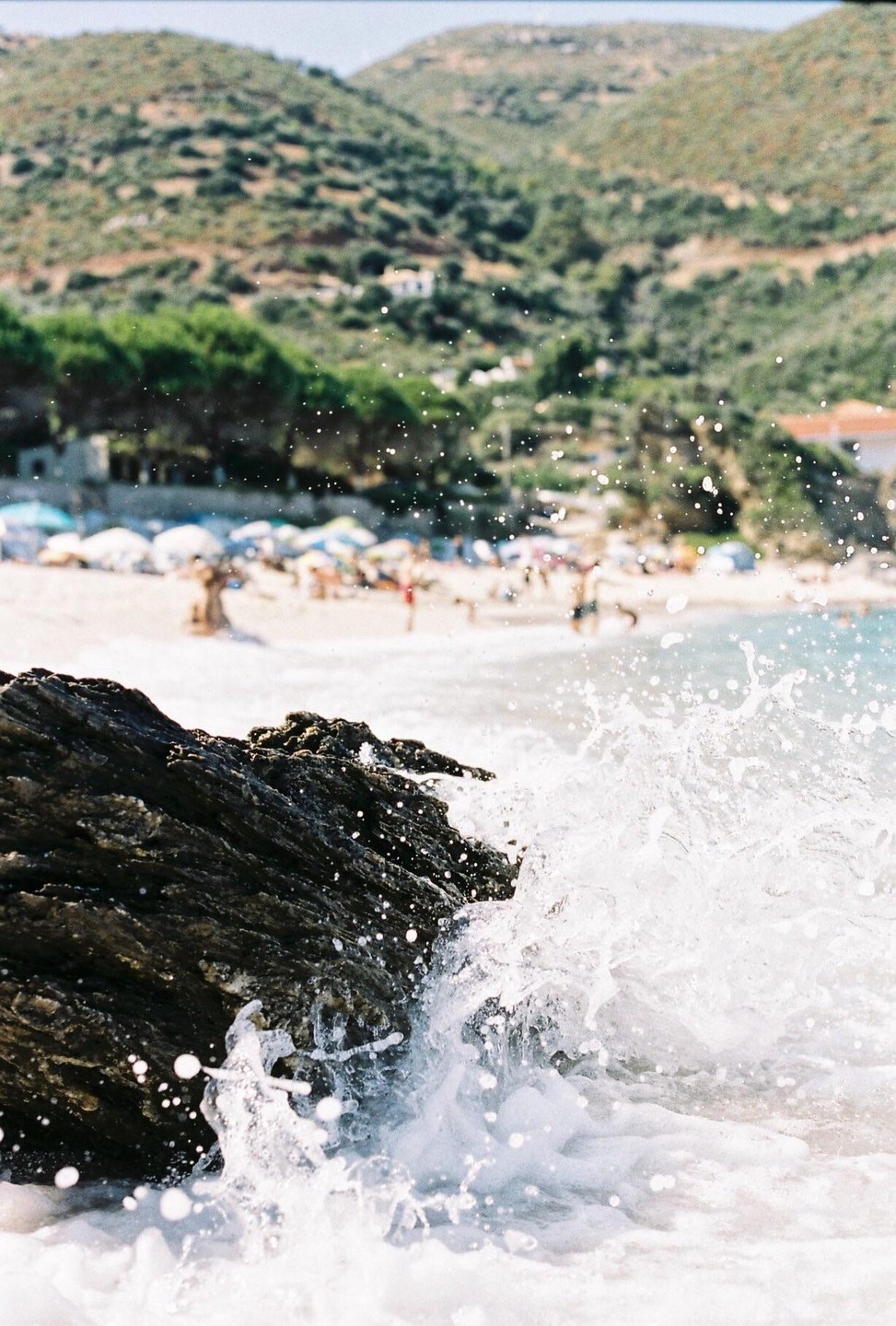A wave crashes against a rock on a beach with mountains in the background.