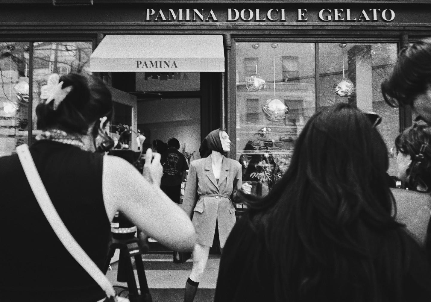 A woman in a blazer poses in front of a storefront with "Pamina Dolci e Gelato" written above it, as other people stand around her.