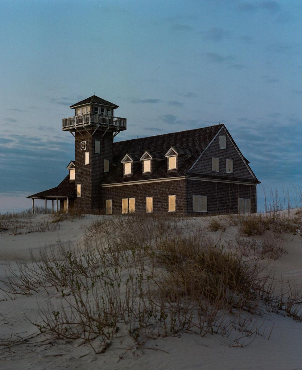 A large wooden house with a tower stands on a sandy dune with sparse vegetation under a twilight sky.