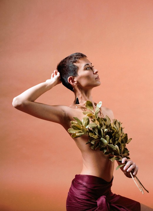 A woman with short hair and a maroon sarong holds a bunch of leaves in front of her chest.