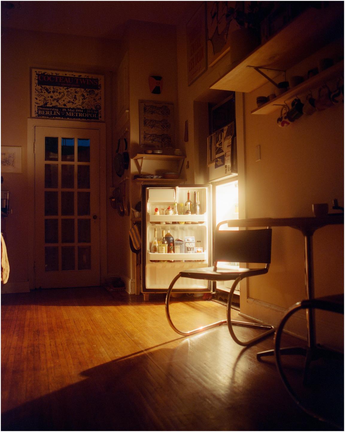 An open refrigerator illuminates a room with a wooden floor and a chair.