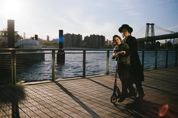 A couple stands on a wooden pier next to a body of water with a bridge and city skyline in the background.