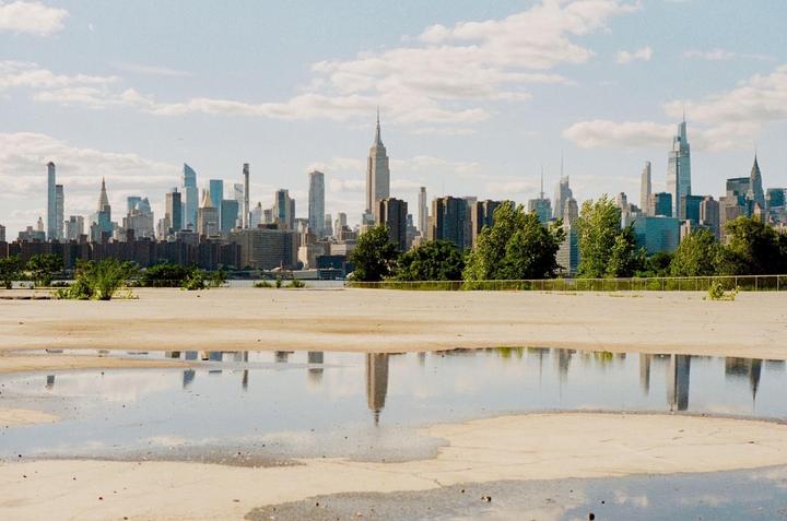 A cityscape with tall buildings is reflected in a puddle on a concrete surface.