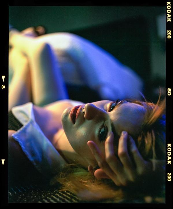 A woman with red lipstick and a nose piercing lies on her back, looking up with her hand near her face.