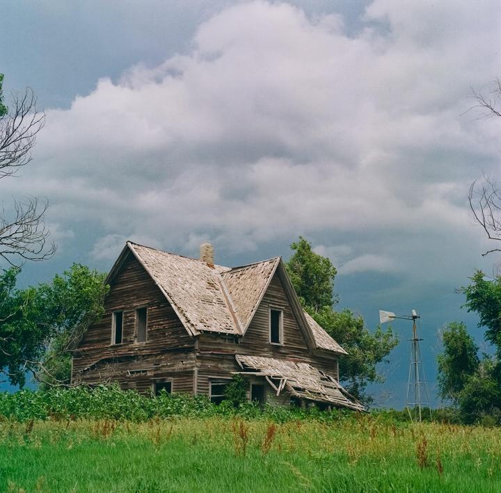An abandoned wooden house with a damaged roof stands in a field under a cloudy sky, with a windmill visible in the distance.