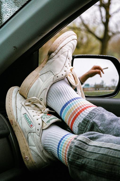 A person's feet in white sneakers and rainbow-striped socks are propped up in a car, with a hand holding a cigarette visible in the side mirror.