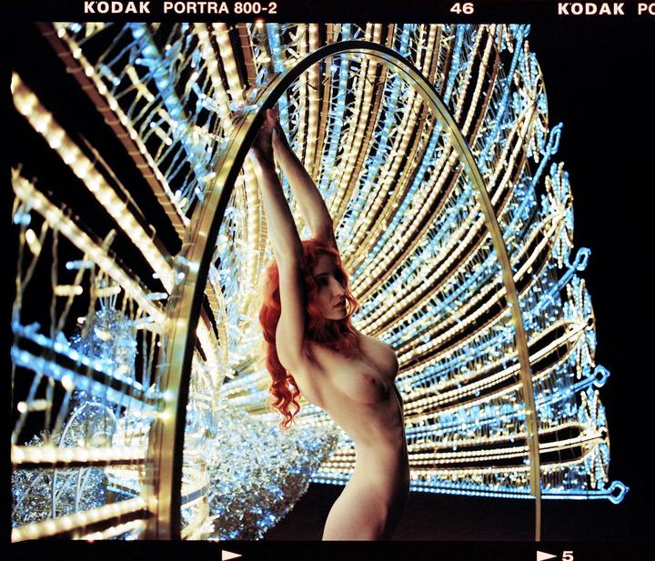 A nude woman with long red hair poses with her arms raised, framed by a large, illuminated peacock sculpture at night.