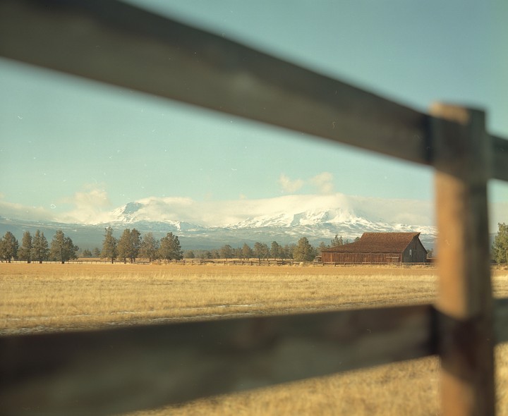 A wooden fence crosses the frame in front of a field with a barn and snow-capped mountains in the distance.