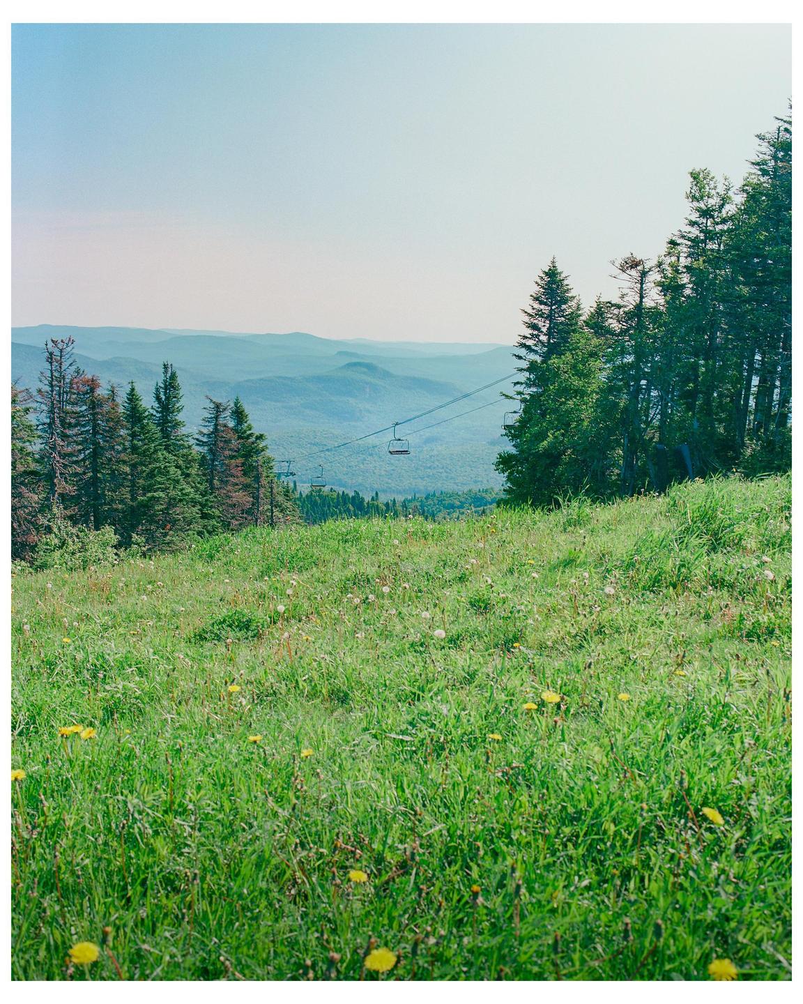 A ski lift is visible above a grassy hill with trees and mountains in the distance.