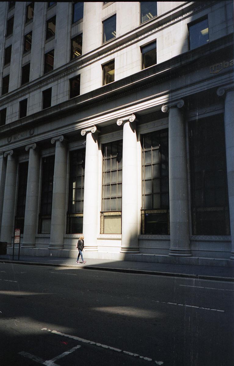 A person walks along a sunlit sidewalk in front of a large building with columns and windows.
