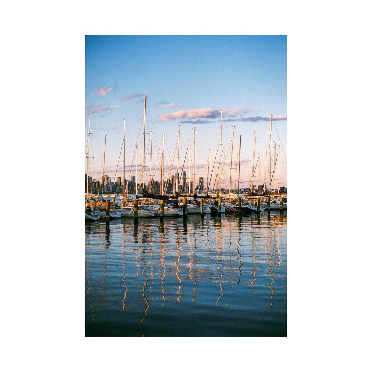 A marina full of sailboats is reflected in the water with a city skyline in the background.