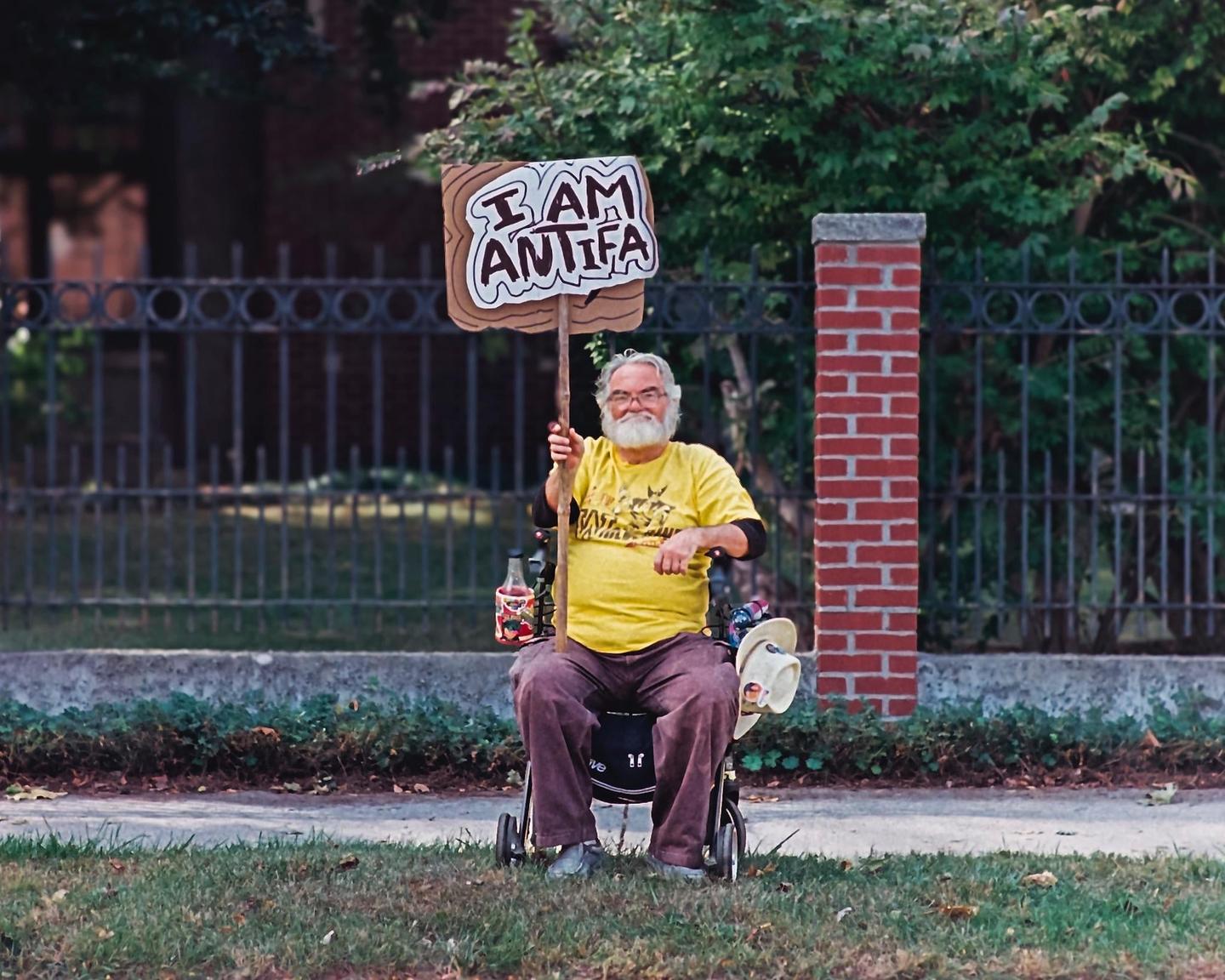 A man in a wheelchair holds a sign that reads "I AM ANTIFA" on a grassy area next to a sidewalk and a fence.