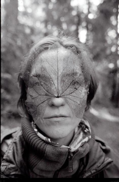 A woman with a large leaf covering her eyes and nose looks directly at the viewer.
