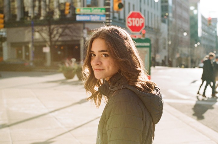 A woman with long hair and a jacket looks over her shoulder on a city street with a stop sign and buildings in the background.