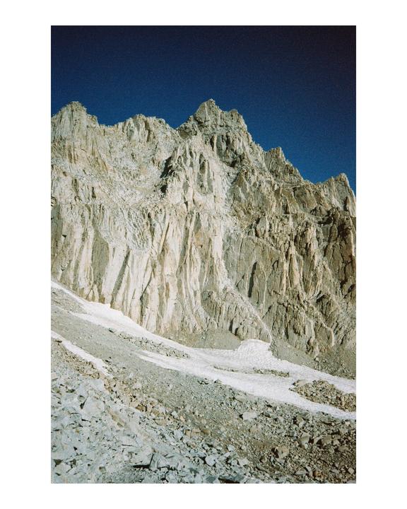 A rugged mountain peak with patches of snow and scree at its base is visible under a clear blue sky.