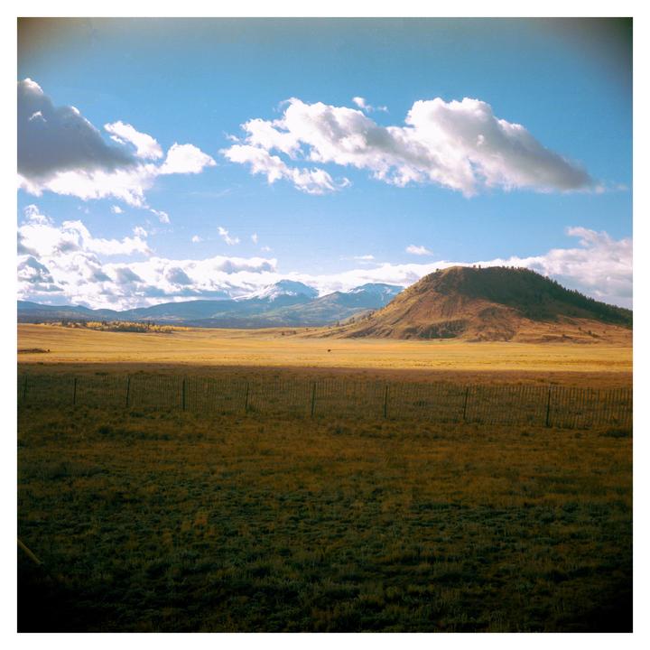 A fence separates a sunlit field from a shadowed field, with mountains and a hill in the background under a cloudy sky.