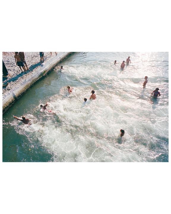 A group of children and adults are swimming and playing in the foamy water next to a sandy bank.
