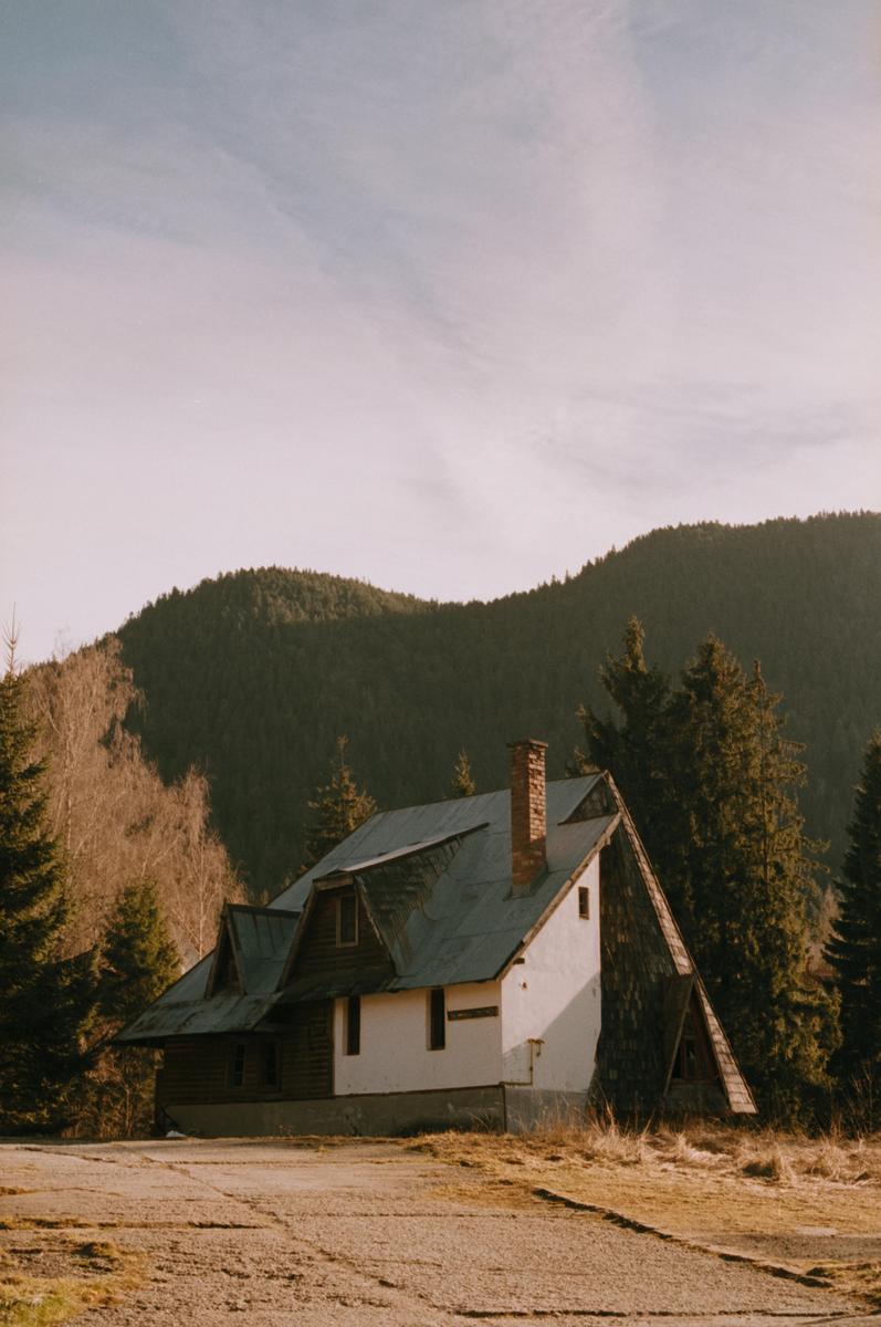 A small house with a chimney stands in a clearing surrounded by trees and mountains.