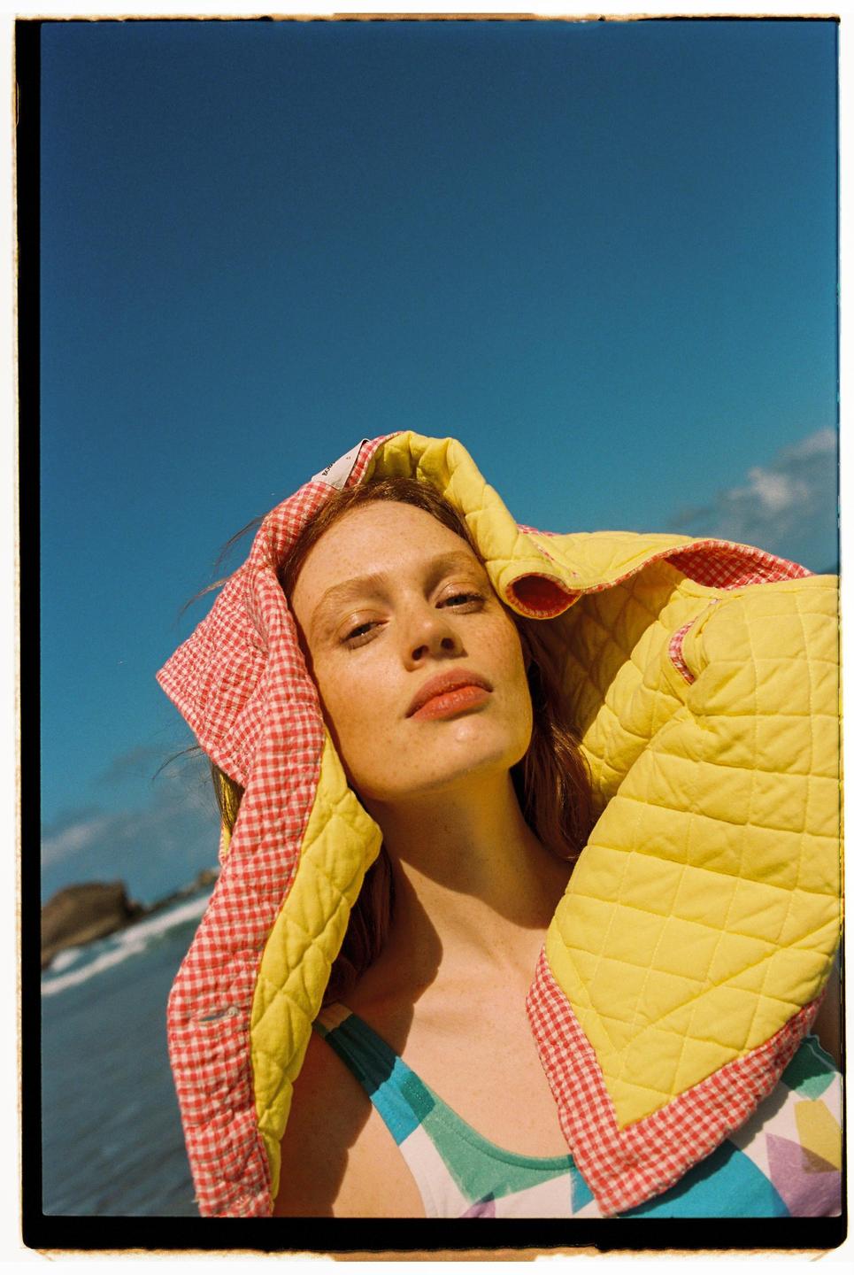 A woman with freckles and red hair looks up at the camera while wearing a colorful swimsuit and a yellow and red checkered quilted blanket over her head.