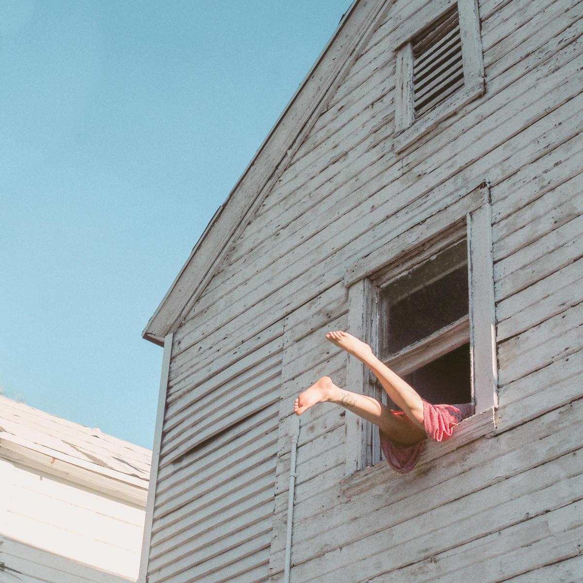 A person's legs extend out of an open window of a white wooden house.