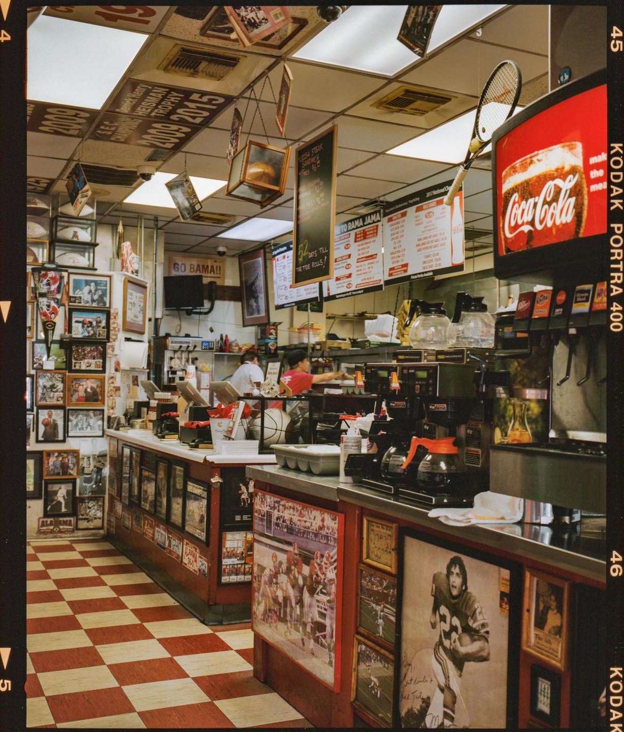Two women work behind the counter of a diner with a checkered floor and sports memorabilia on the walls.