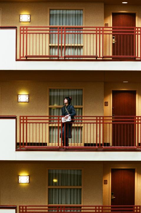 A man stands on a hotel balcony holding a newspaper, looking up.
