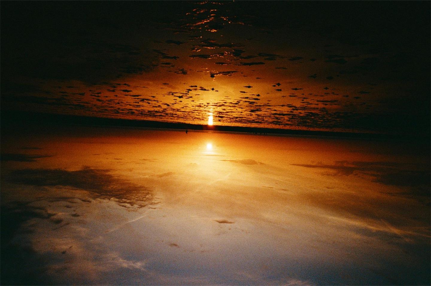 The sun sets over a body of water, reflecting on the surface and illuminating the clouds above.