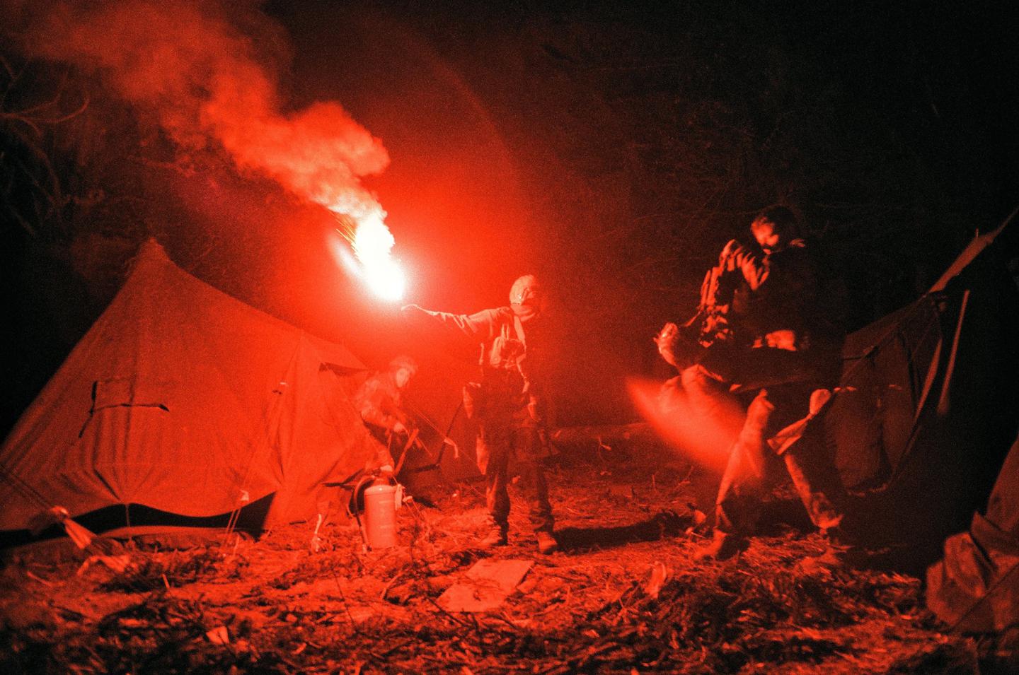 Three men are gathered around a red flare at night, with two tents visible in the background.