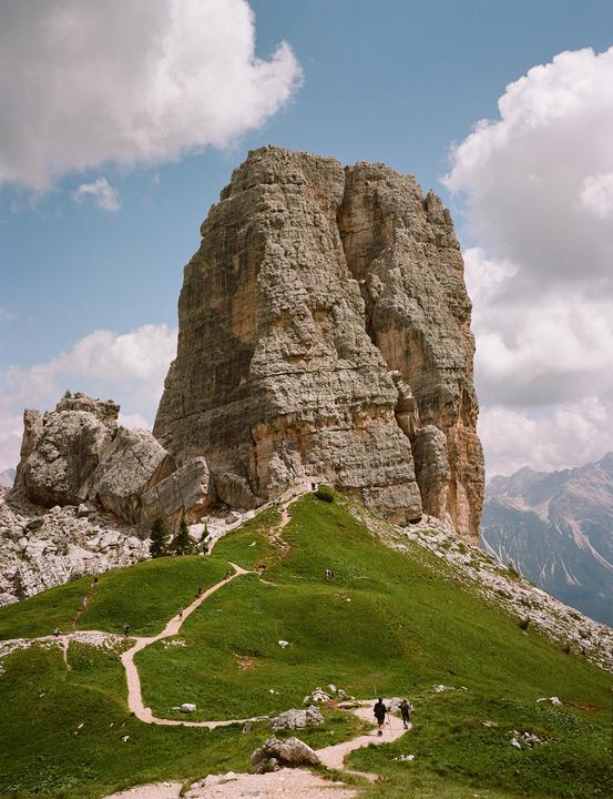 A winding dirt path leads up a grassy hill towards a towering rock formation under a cloudy sky.