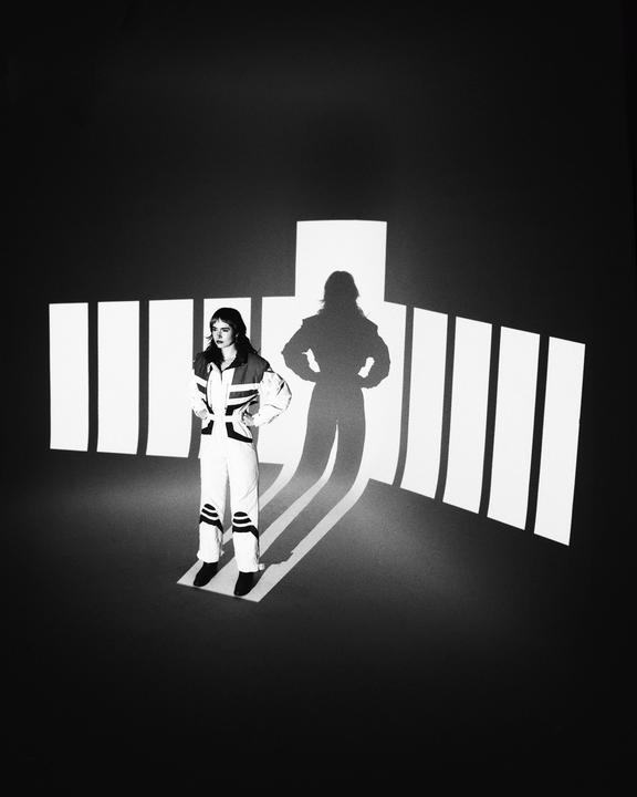 A woman in a jumpsuit stands with her hands on her hips, casting a long shadow against a backdrop of projected light rectangles.