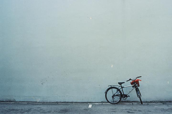 A bicycle with a red basket is parked against a light blue wall on an asphalt street.