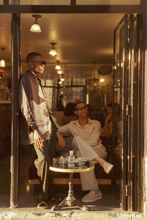 Two men are relaxing at a cafe table with drinks, one standing and one sitting, with other patrons visible in the background.