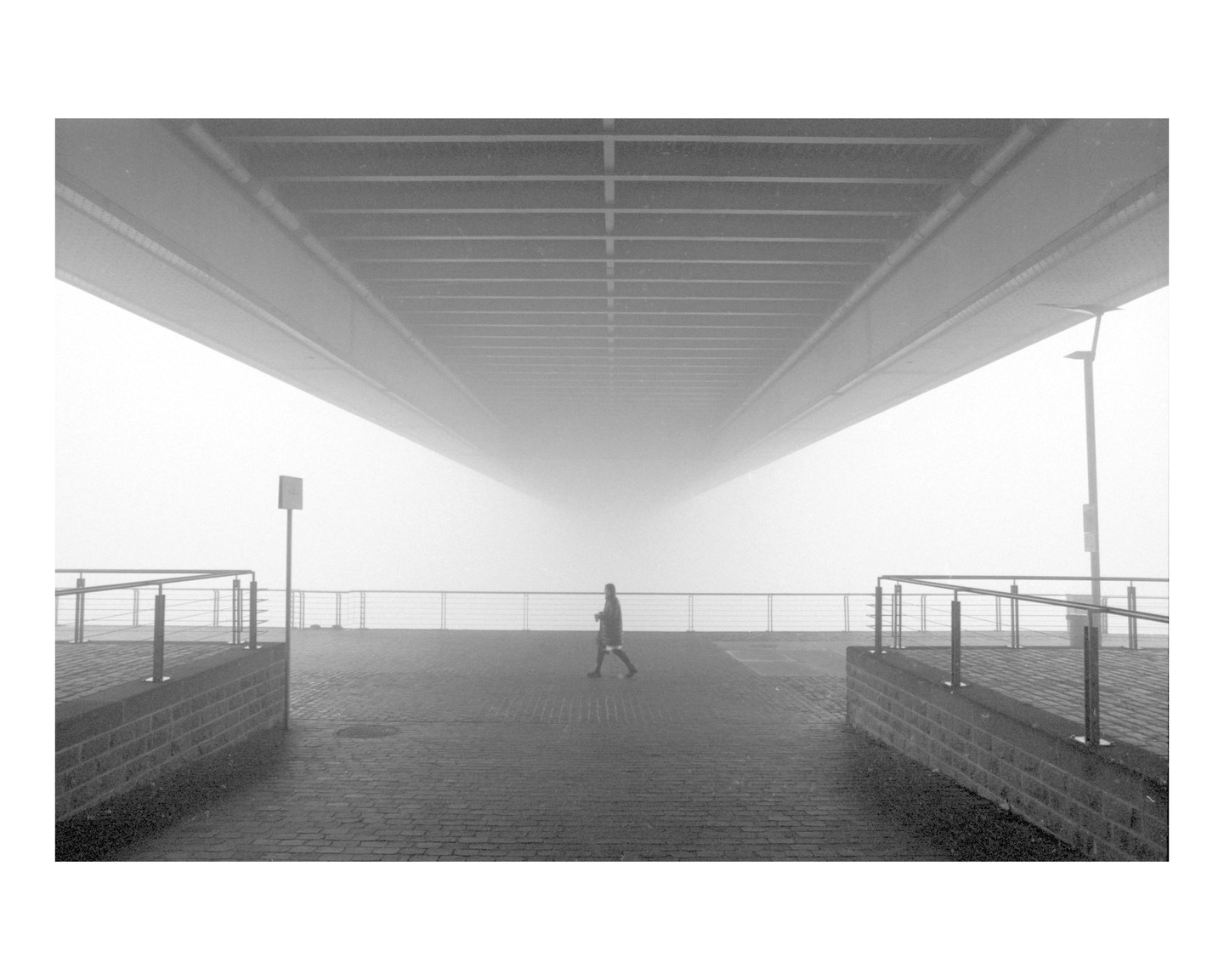 A person walks on a paved path under a bridge on a foggy day.