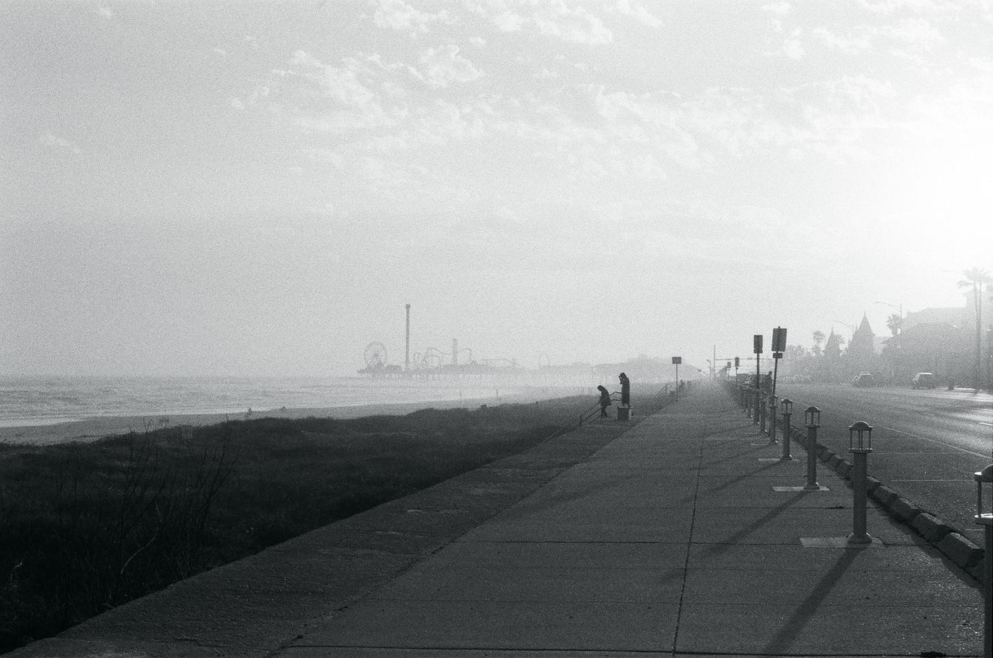 Two people stand on a boardwalk next to a beach with an amusement park in the distance.