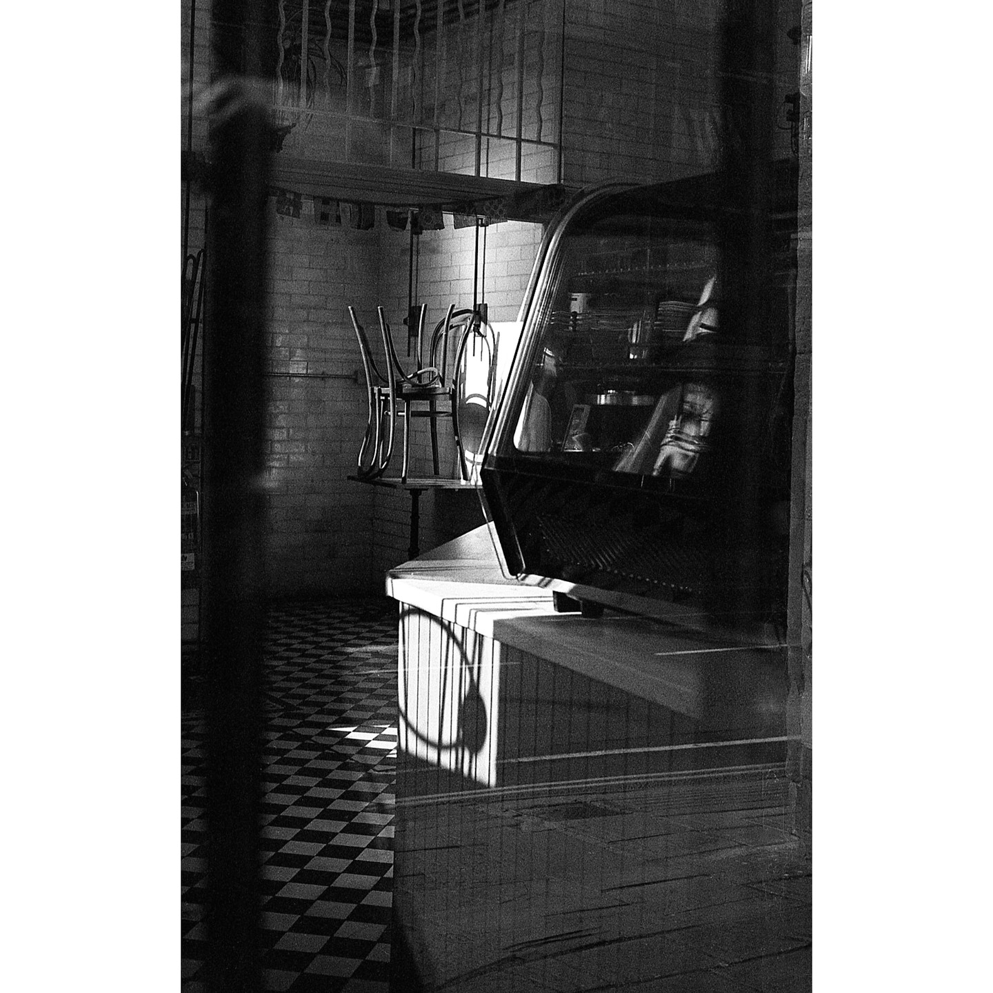 Sunlight casts shadows on a checkered floor and a counter with a display case, with chairs stacked on a table in the background.