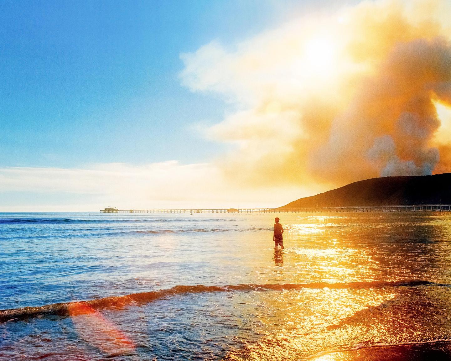 A person stands in the ocean with a pier in the distance and a large plume of smoke rising from behind a hill.