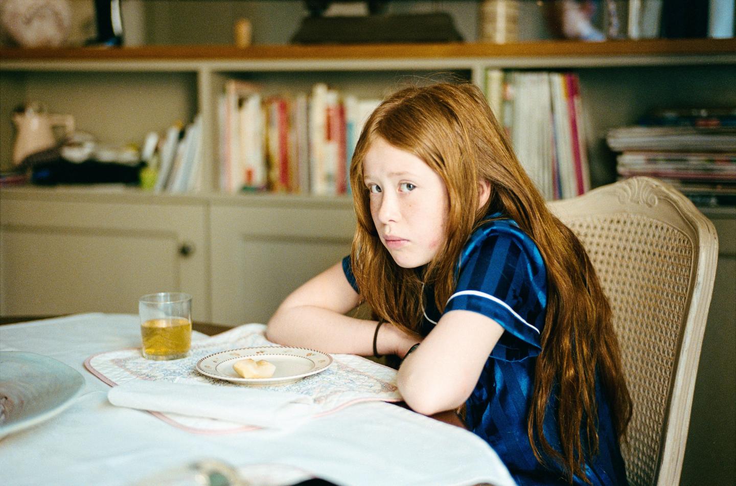 A girl with long red hair sits at a table with a glass of juice and a small pastry.