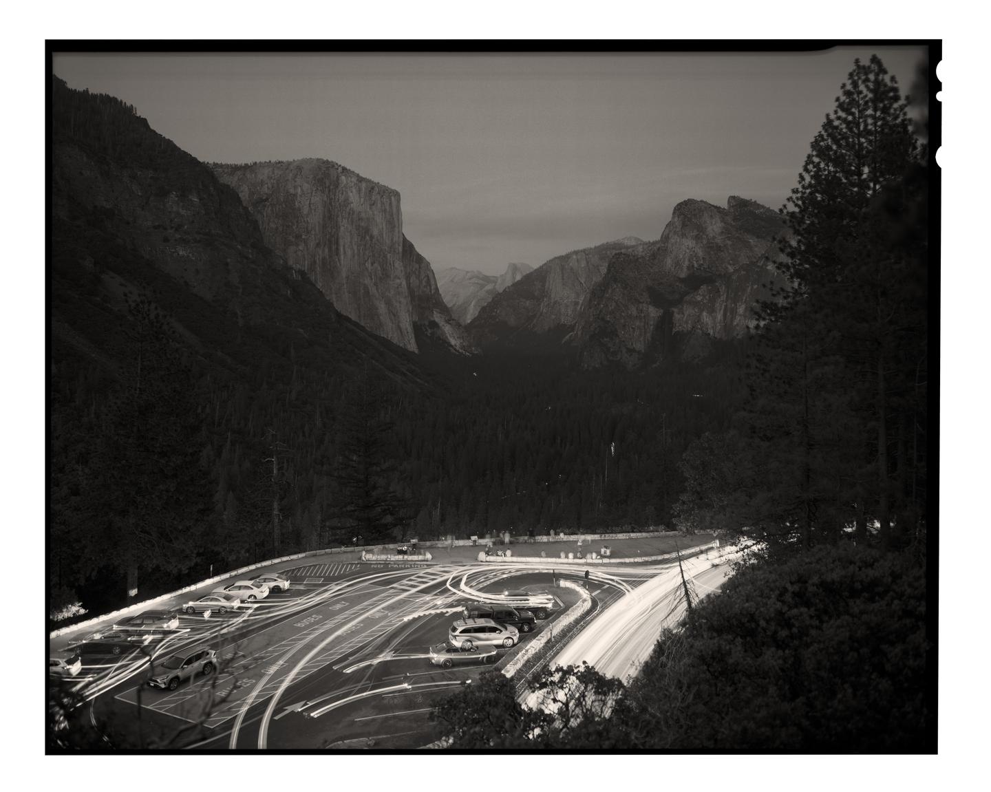 A black and white landscape shows a valley with mountains and a forest, with a road and parking lot in the foreground.