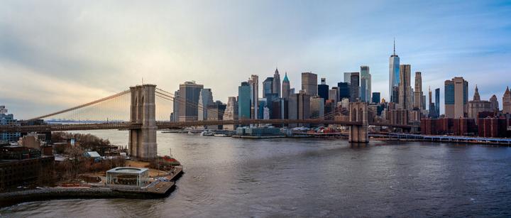 A panoramic view shows the Brooklyn Bridge spanning a river with the New York City skyline in the background.