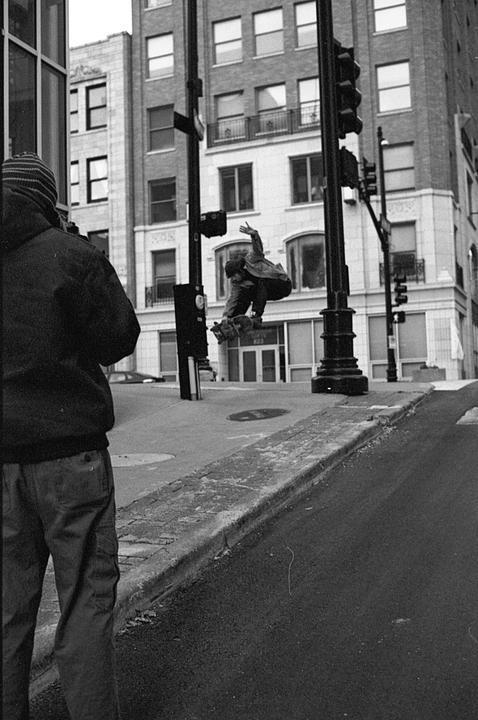 A person in a striped hat and jacket stands on a sidewalk watching a skateboarder in mid-air.