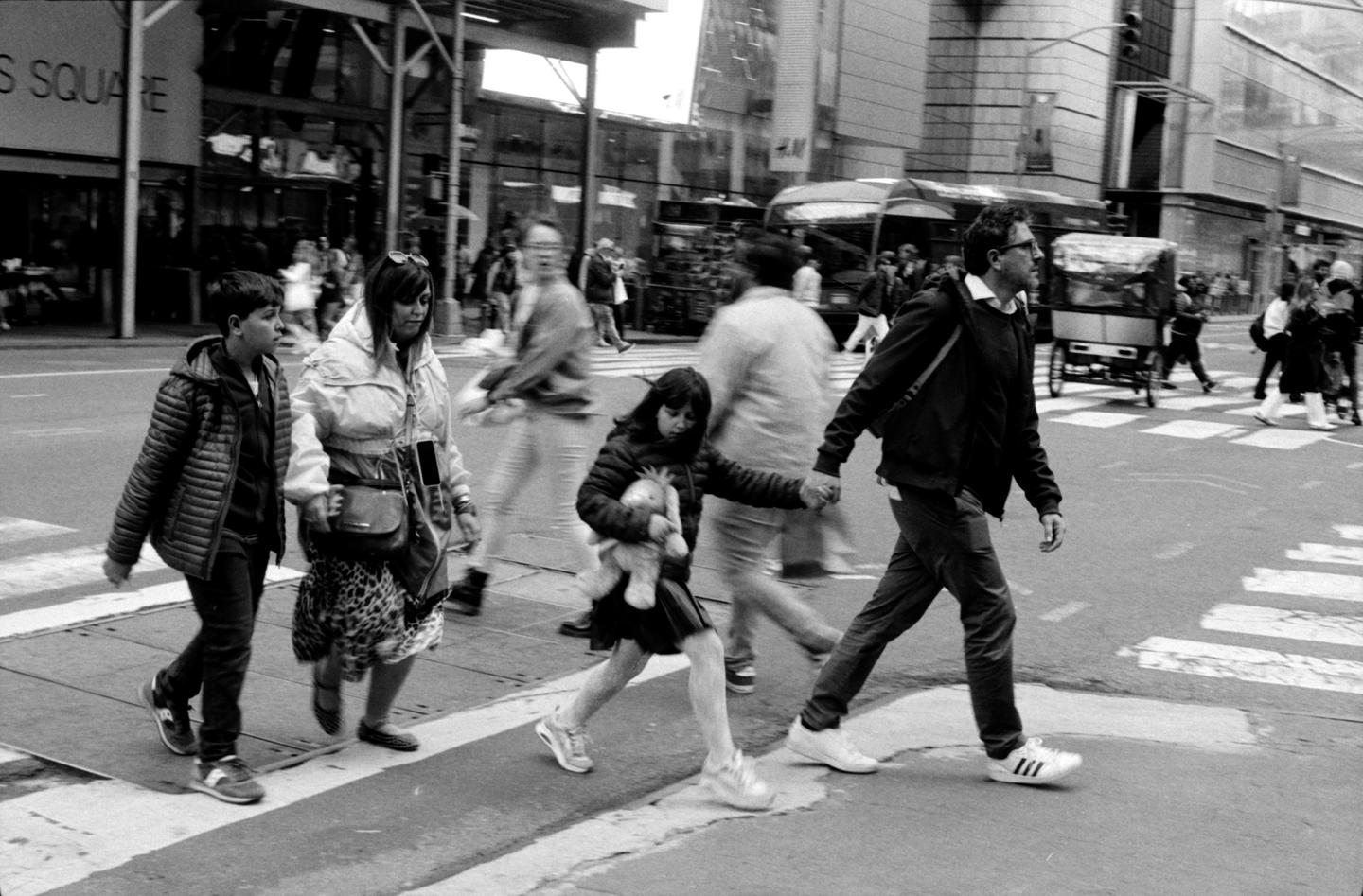 A family crosses a street in a busy city, with a young girl holding a stuffed animal.