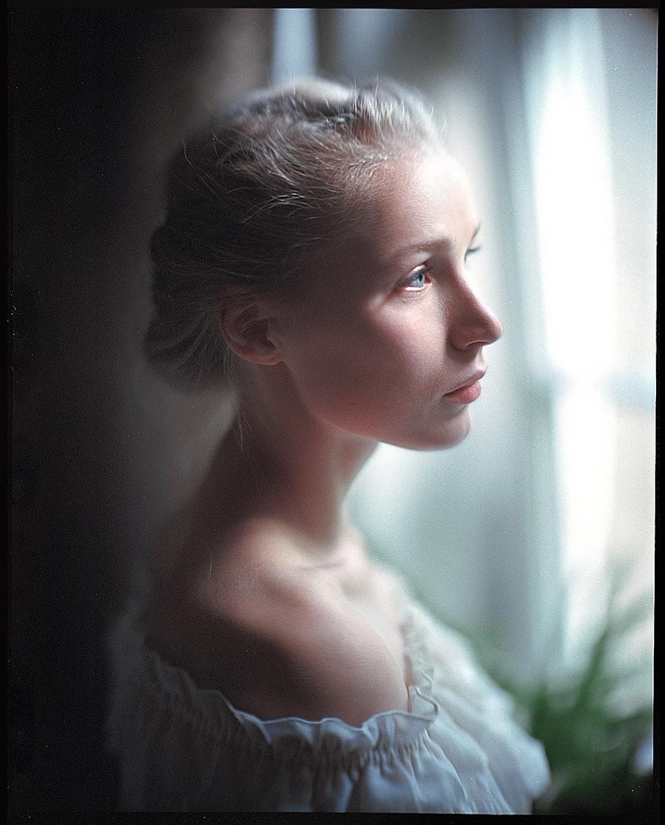 A woman with blonde hair looks out a window, illuminated by soft light.