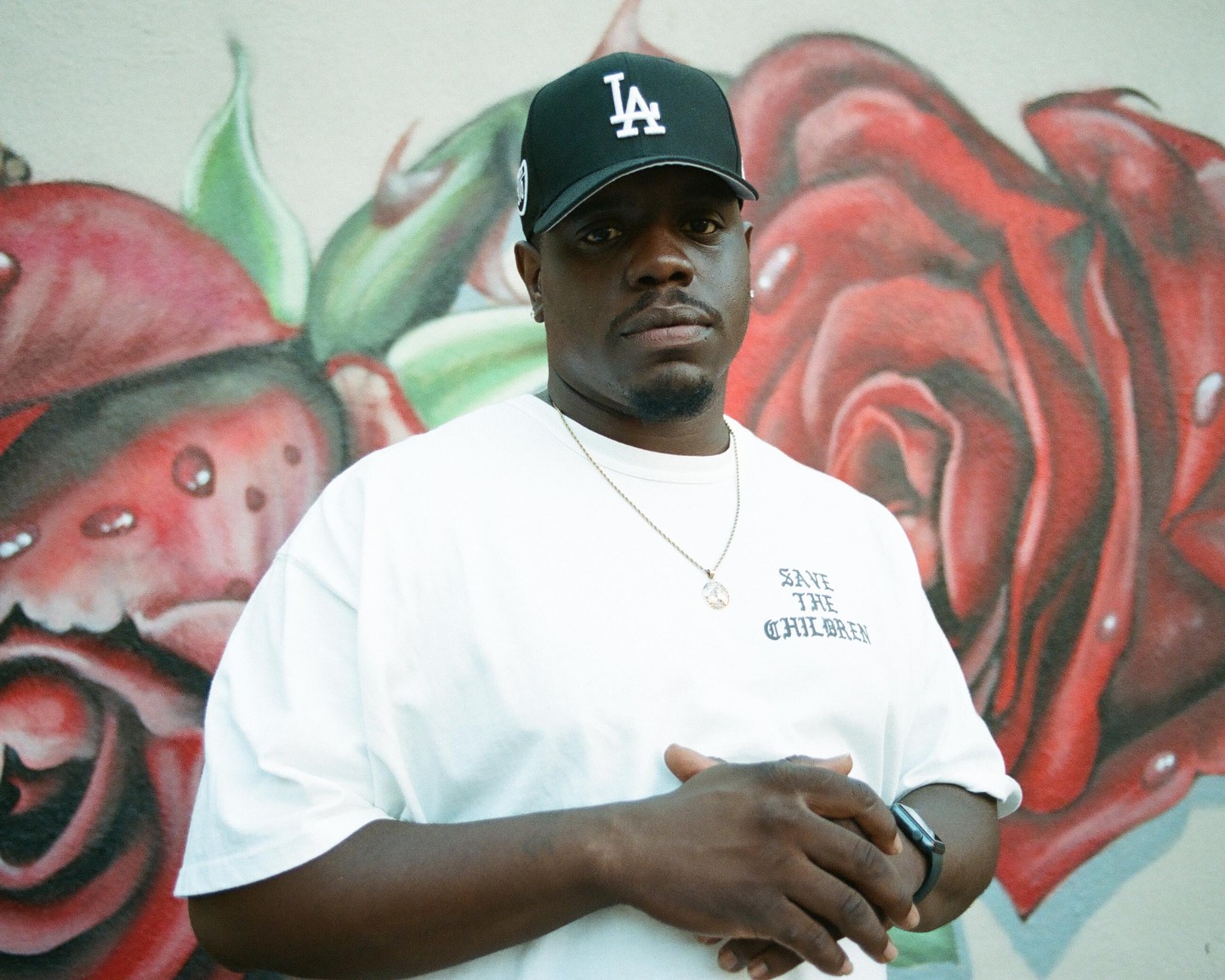 A man wearing a baseball cap and a white t-shirt stands in front of a wall with red rose graffiti.