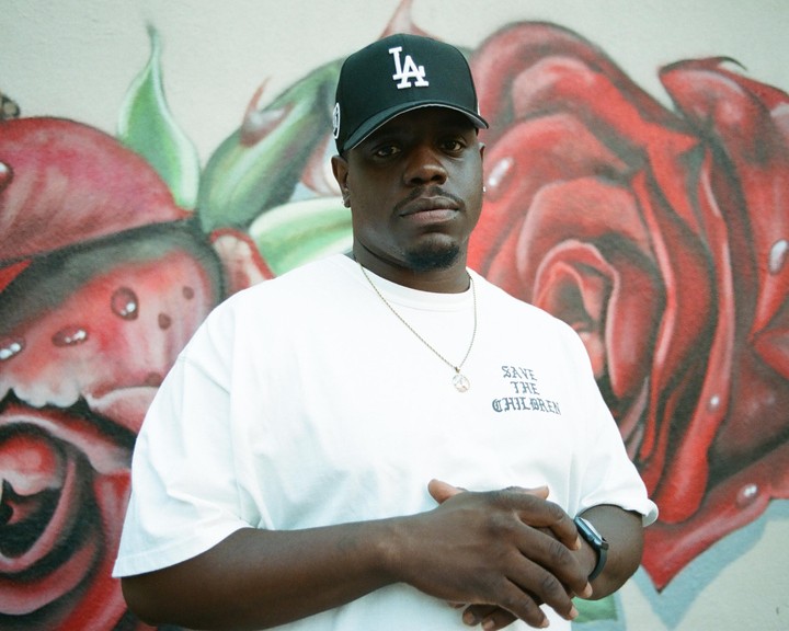 A man wearing a baseball cap and a white t-shirt stands in front of a wall with red rose graffiti.