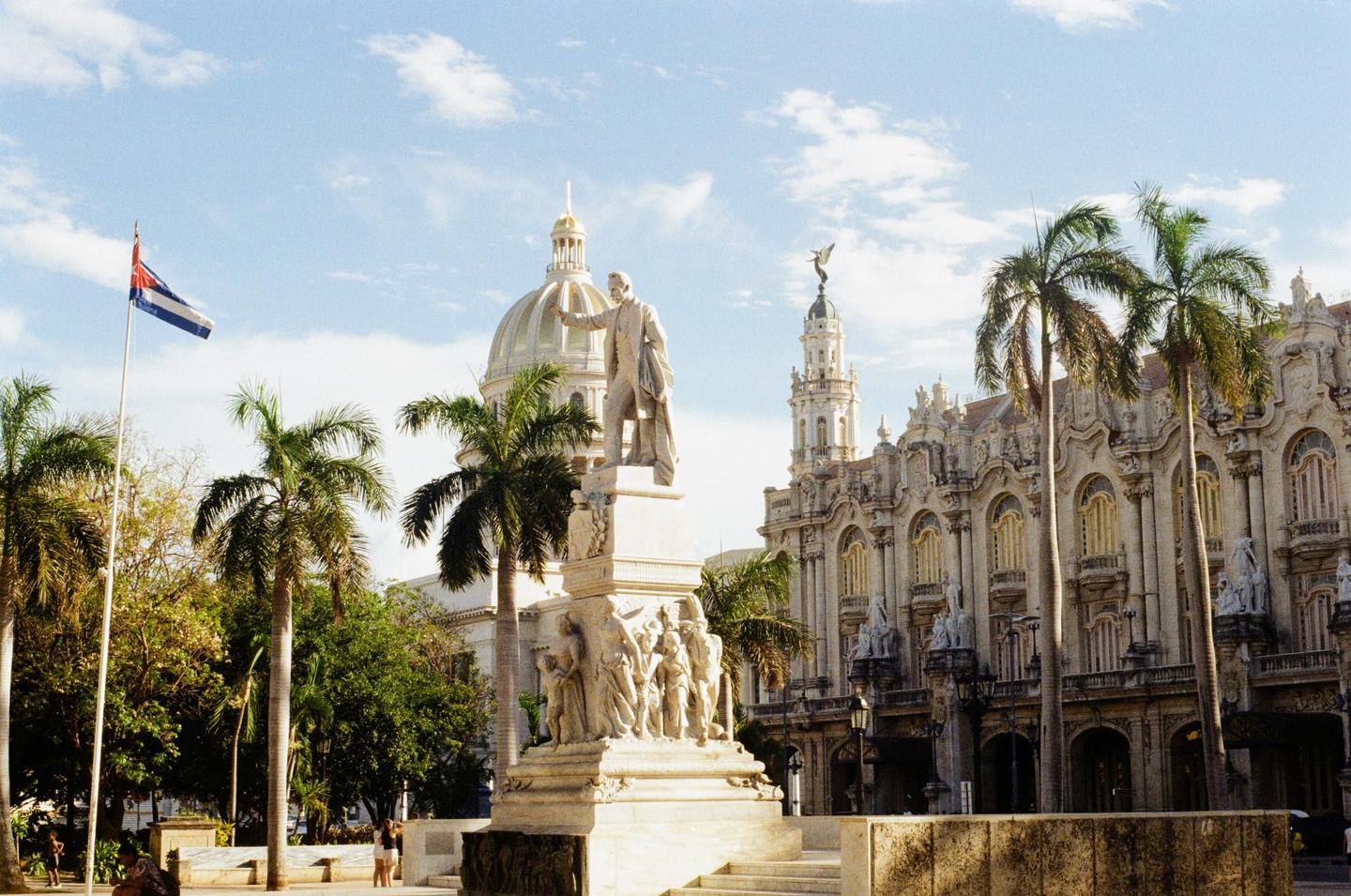 A statue of a man stands in front of a domed building and a large ornate building with palm trees and a Cuban flag.