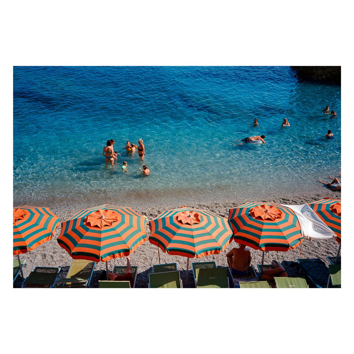 A group of people swim in clear blue water next to a beach lined with striped umbrellas and lounge chairs.