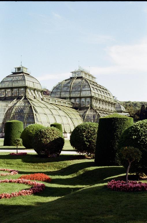 Two large glass greenhouses are visible behind manicured hedges and flowerbeds in a garden.