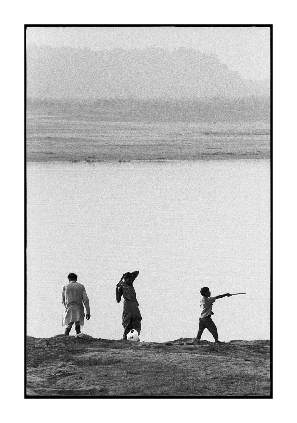 Three people stand on a riverbank with a mountain in the distance.
