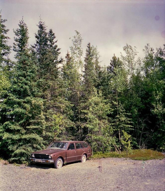 A brown station wagon is parked on a gravel lot next to a dense forest.