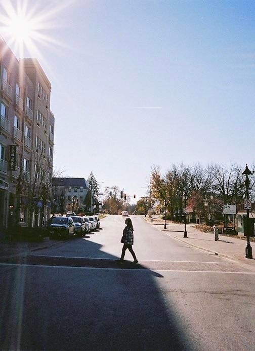 A woman crosses a street with parked cars and buildings under a bright sun.
